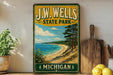 A vintage-style sign for J.W. Wells State Park in Michigan, featuring a scenic beach landscape with pine trees and a blue sky.