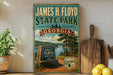A vintage-style metal sign for James H. Floyd State Park in Georgia, featuring a scenic landscape with pine trees, a lake, and a sign that says "James H. Floyd State Park".