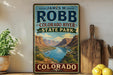 A vintage-style sign for James M. Robb Colorado River State Park, featuring a scenic mountain landscape with a river and a small boat.