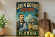 A vintage-style metal sign for the John Gorrie Museum in Florida, featuring an illustration of a man in a suit and tie standing next to a large ice machine.