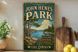 A vintage-style metal sign for John Hens Park in Wisconsin, featuring a scenic landscape with a lake and pine trees.