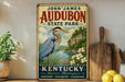 A vintage-style sign for John James Audubon State Park in Kentucky, featuring an illustration of a heron in a grassy landscape with a river and mountains in the background.