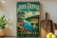 A vintage-style sign for John Tanner State Park in Georgia, featuring a scenic landscape with pine trees, a lake, and mountains.