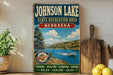 A vintage-style sign for Johnson Lake, Nebraska, featuring a scenic image of a lake surrounded by mountains and a boat on the water.