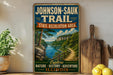 A vintage-style sign for the Johnson-Sauk Trail, featuring a scenic illustration of a river and mountains, is displayed on a wooden shelf next to a potted plant and a cutting board.