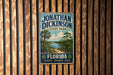 A vintage-style metal sign for Jonathan Dickinson State Park, featuring a scenic illustration of a beach and palm trees, mounted on a wooden wall.