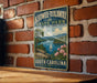 A vintage-style metal sign for Keowee-Toxaway State Park in South Carolina, featuring a scenic landscape with a lake, mountains, and a sailboat.
