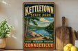 A vintage-style sign for kettletown state park, featuring a scenic landscape with a lake and mountains, is displayed on a wooden shelf next to a potted plant and a cutting board.