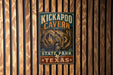 A metal sign with the text "KICKAPOO CAVEN" and "STATE PARK TEXAS" is displayed on a wooden wall.