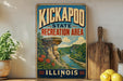 A vintage-style sign for the Kickapoo State Recreation Area in Illinois, featuring a scenic landscape with a river, cliffs, and flowers.