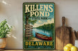 A vintage-style sign for Killens Pond State Park in Delaware, featuring a canoe and a scenic landscape with trees and a lake.