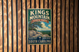 A vintage metal sign for Kings Mountain State Park in South Carolina, featuring a scenic landscape with a large rock formation and a palm tree.