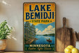 A vintage-style sign for Lake Bemidji State Park in Minnesota, featuring a scenic illustration of a lake surrounded by pine trees and a person in a canoe.