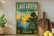 A vintage-style sign for Lake Griffin State Park in Florida, featuring a seagull, palm trees, and a sunset over the water.