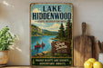 A vintage-style metal sign for Lake Hiddenwood, South Dakota, featuring a scenic illustration of a lake surrounded by mountains and pine trees.