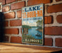 A vintage-style sign for Lake Le-Aqua-NA, a state recreation area in Illinois, featuring a scenic image of a lake with boats and people fishing.