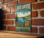 A vintage-style sign for Lake Louise State Park in Minnesota, featuring a scenic landscape with pine trees, a lake, and mountains.
