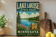A vintage-style sign for Lake Louise State Park in Minnesota, featuring a scenic landscape with pine trees, a lake, and mountains.
