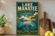 A vintage-style sign for Lake Manatee State Park in Florida, featuring a manatee swimming in the water and surrounded by palm trees and other natural scenery.