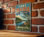 A vintage-style metal sign for Lake Maria State Park in Minnesota, featuring a scenic landscape with a lake, mountains, and pine trees.