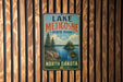 A vintage metal sign for Lake Metigoshe State Park in North Dakota, featuring a scenic landscape with pine trees, a lake, and mountains.