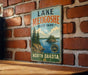 A vintage-style metal sign for Lake Metigoshe State Park in North Dakota, featuring a scenic landscape with pine trees and a lake.