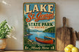 A vintage-style sign for Lake St. George State Park in Maine, featuring a scenic illustration of a lake, mountains, and a canoe.