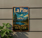 A metal sign with a scenic mountain landscape and water, mounted on a wall next to a plant.