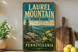 A vintage-style metal sign for Laurel Mountain State Park in Pennsylvania, featuring a scenic mountain landscape with pine trees and flowers.