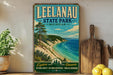 A vintage-style metal sign for Leelau State Park, Michigan, featuring a scenic coastal landscape with pine trees, a beach, and a lake.