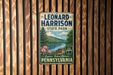 A vintage-style metal sign for Leonard Harrison State Park in Pennsylvania, featuring a scenic landscape with a river, trees, and mountains.