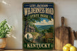 A vintage-style sign for the Levi Jackson Wilderness Road in State Park, Kentucky, featuring a scenic image of a forest and a cabin.