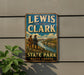 A vintage-style metal sign for Lewis and Clark State Park in North Dakota, featuring a scenic landscape with two figures standing on a cliff overlooking a river.