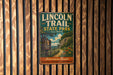 A vintage metal sign for the Lincoln Trail State Park in Illinois, featuring a scenic illustration of a trail surrounded by nature.
