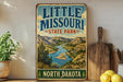 A vintage-style metal sign for Little Missouria State Park in North Dakota, featuring a scenic landscape with mountains, a river, and a wooden paddle.