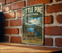 A vintage-style metal sign for Little Pine State Park in Pennsylvania, featuring a scenic landscape with pine trees, a lake, and mountains.