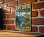 A vintage-style metal sign for Lost Creek State Park in Montana, featuring a scenic landscape with mountains, a river, and pine trees.