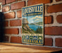 A vintage-style metal sign for the Louisville State Recreation Area in Nebraska, featuring a scenic landscape with mountains, water, and camping symbols.