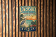 A vintage-style metal sign for "Lovers Key State Park" in Florida, featuring a beach scene with palm trees, a crane, and a sunset.