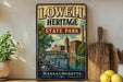 A vintage-style metal sign for Lowell Heritage State Park, featuring a scenic illustration of a river and buildings, is displayed on a shelf next to a wooden cutting board and some lemons.