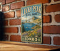 A vintage-style metal sign for Lucky Peak State Park in Idaho, featuring a scenic mountain landscape with a lake and surrounding vegetation.