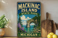 A vintage-style sign for Mackinac Island State Park in Michigan, featuring a scenic landscape with a rock arch, pine trees, and a lake.