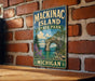 A vintage-style metal sign for Mackinac Island State Park in Michigan, featuring a scenic landscape with a bridge, water, and trees.
