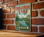 A vintage metal sign for Madison Lake State Park in Ohio, featuring a scenic illustration of a lake with a sailboat and surrounding landscape.