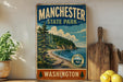 A vintage-style metal sign for Manchester State Park in Washington, featuring a scenic landscape with pine trees, a beach, and a mountain range.