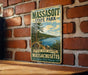 A vintage metal sign for Massasoit State Park, featuring a scenic landscape with pine trees, a lake, and mountains.