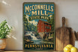 A vintage-style sign for McConnells Mill State Park in Pennsylvania, featuring a red brick building with a watermill and a waterfall.