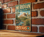A vintage-style metal sign for Meridian State Park in Texas, featuring a scenic landscape with mountains, a river, and desert vegetation.
