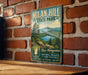 A vintage metal sign for Milan Hill State Park, featuring a scenic mountain landscape and a lake, is displayed on a brick wall.