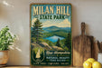 A vintage-style metal sign for Milan Hill State Park in New Hampshire, featuring a scenic landscape with mountains, a lake, and pine trees.